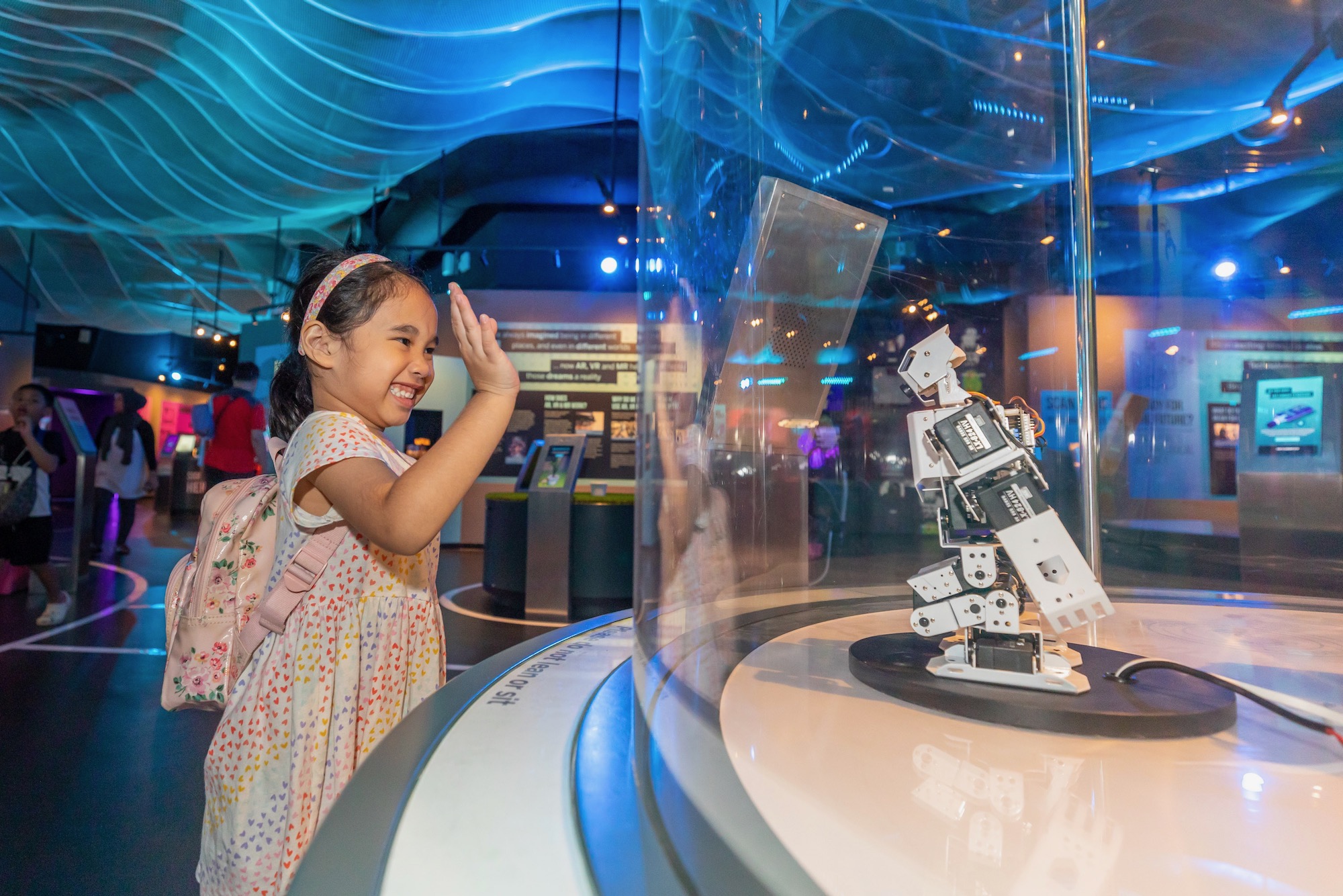 A girl waving to a robot enclosed in a glass case